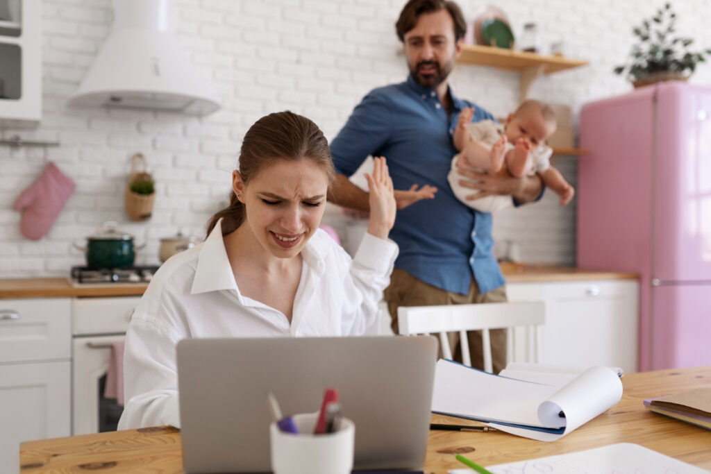 Donna in smart working stressata al computer in cucina ignora marito e figlio neonato, esempio di mancato diritto alla disconnessione e burnout familiare.