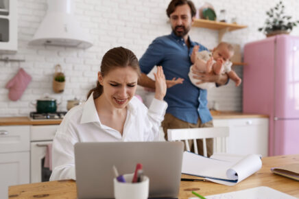 Donna in smart working stressata al computer in cucina ignora marito e figlio neonato, esempio di mancato diritto alla disconnessione e burnout familiare.
