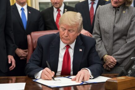 Donald Trump seduto alla scrivania mentre firma un documento ufficiale, circondato da collaboratori.