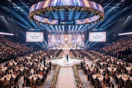 Foto panoramica dell'interno dell'Unipol Dome allestito per un matrimonio sfarzoso: sposi al centro su una pista circolare decorata di fiori, circondati da tavoli rotondi per la cena e spalti affollati sotto luci d'atmosfera.